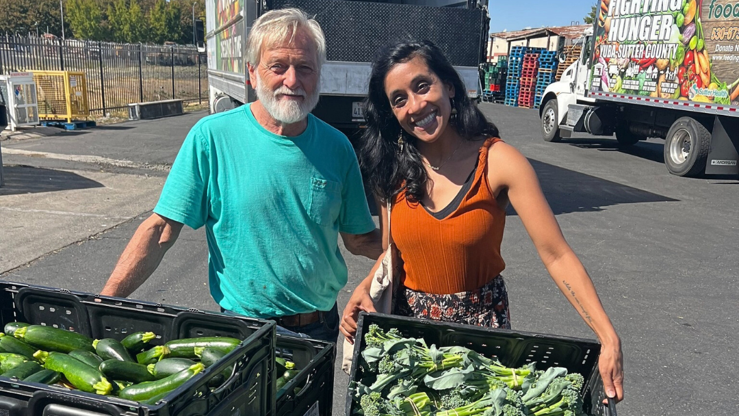 A man and women smiling while holding crates of green vegetables in front of two large food distribution trucks.