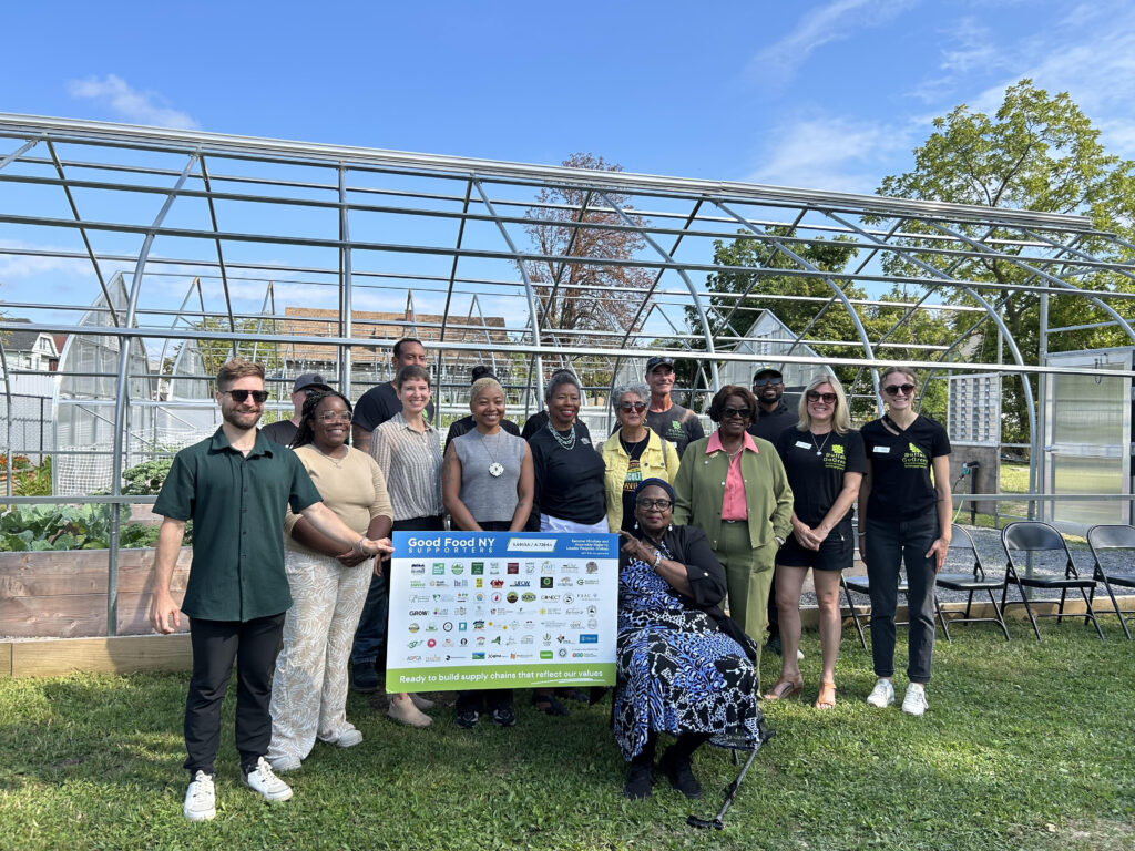 A group of 15 individuals smiling and convening around a sign listing many organizations' names in front of a greenhouse structure.