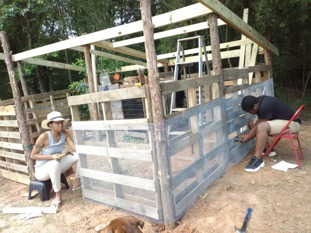 Two individuals sitting in chairs on opposing sides of a large wooden chicken coop, hammering chicken fencing to the wood.